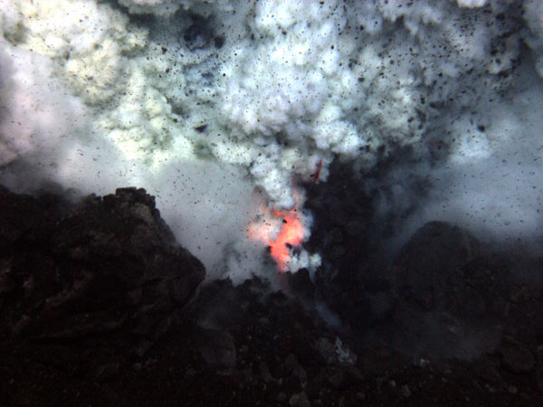 An explosion near the summit of West Mata volcano throws ash and rock, and molten lava glows below. 