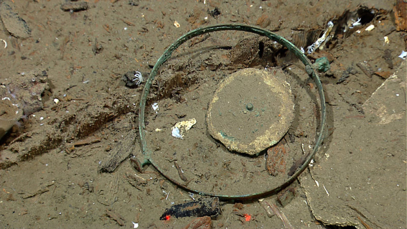 Several pieces of the ship’s compass were found in the aft part of the vessel. The compass card inside the gimbaled mount, shown here, is in an area where the ship’s binnacle would have been located. The two red dots at the bottom of the picture are laser lights that come from the ROV Little Hercules. ROVs often mount two parallel lasers to take measurements and these laser lights are 10 centimeters apart. Image courtesy NOAA Okeanos Explorer Program.