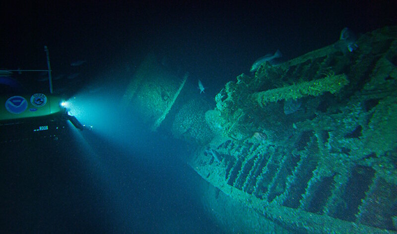 A submersible shines its lights on the wreck of U-576 lying on its starboard side, showing the submarine's conning tower and the deck gun in the foreground.