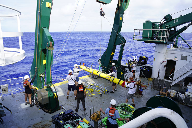 A REMUS 600 vehicle is launched from the A-frame of Research Vessel Kilo Moana during the Deepwater Surveys of World War II U.S. Cultural Assets in the Saipan Channel expedition. Crew and staff wear hard hats, work vests, and steel-toed boots during deck operations for safety. The A-frame lifts the vehicle using a strap attached to a brailer, which releases the vehicle once in the water. A red line is attached to the nose of the vehicle and is released remotely when the vehicle is ready to dive.