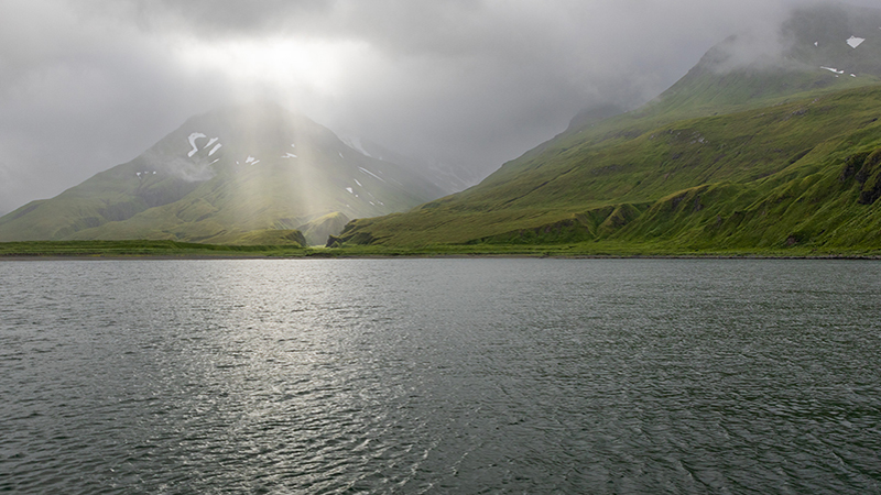 Serene landscape with a body of water, green hills, and cloudy mountains with sunlight rays.