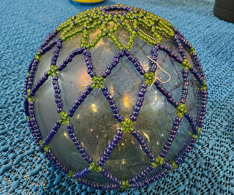 The image shows a spherical object covered with a decorative net made of beads. The sphere appears to be a clear, glass-like material. The beaded net design features a lattice pattern, with blue and green beads arranged in intersecting diagonal lines. The blue beads form the primary structure, while the smaller green beads are placed at the intersections, creating a repeating geometric pattern. The surface beneath the sphere is covered with a textured blue fabric, complementing the colors of the beads.