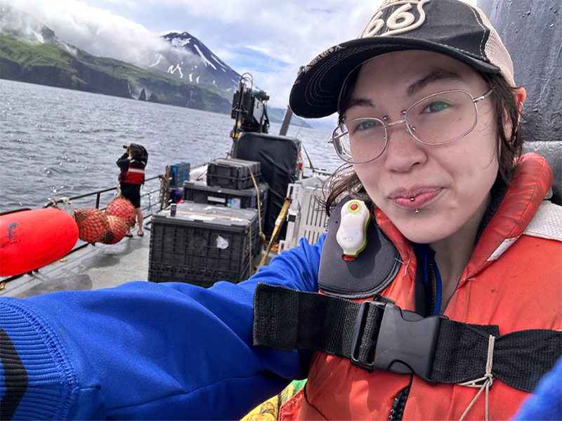 The image shows a person taking a selfie on a boat, with a mountainous landscape and ocean in the background. The person is wearing a hat with the number 66, glasses, a blue jacket, and an orange life vest. They also have a dark lip piercing. Behind them on the boat are crates and red flotation devices. Another person stands farther back, wearing a life jacket and looking towards the water. The background features a snow-capped mountain partially obscured by clouds, with green cliffs along the coastline.