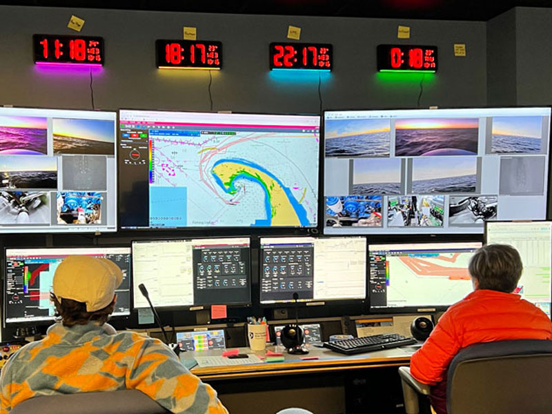 Two people in a control room with multiple monitors displaying maps, images, and data. Digital clocks are mounted above the screens.