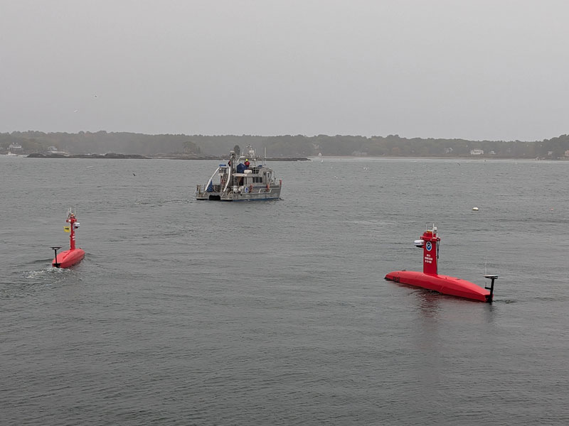 Two red uncrewed surface vehicles on calm water, with a larger ship in the background under a gray sky.