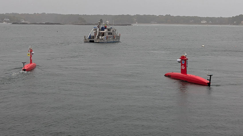 Two red uncrewed surface vehicles on calm water, with a larger ship in the background under a gray sky.