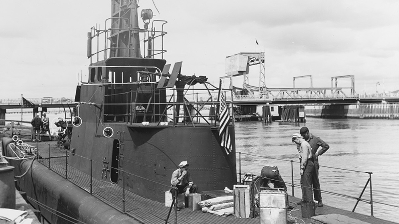 Submarine docked with sailors on deck and a bridge in the background.