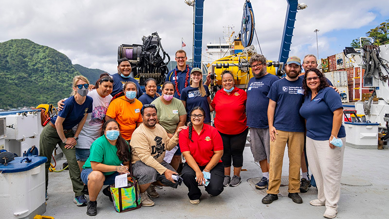 The image depicts a group of sixteen people posing together on the deck of a ship. They are standing or crouching in front of various pieces of marine equipment, such as a large cable reel and mechanical apparatus in the background. The group is diverse in age and apparently gender, with individuals wearing casual clothing including t-shirts, jeans, and shorts. Some are wearing masks. The background features a mountainous, lush green landscape under a cloudy sky, suggesting a tropical location. The ship's deck is equipped with containers and other maritime equipment.