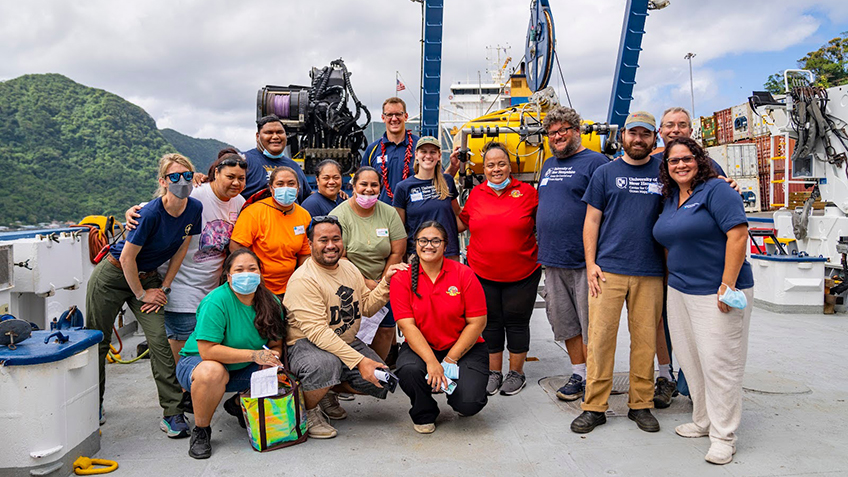 The image depicts a group of sixteen people posing together on the deck of a ship. They are standing or crouching in front of various pieces of marine equipment, such as a large cable reel and mechanical apparatus in the background. The group is diverse in age and apparently gender, with individuals wearing casual clothing including t-shirts, jeans, and shorts. Some are wearing masks. The background features a mountainous, lush green landscape under a cloudy sky, suggesting a tropical location. The ship's deck is equipped with containers and other maritime equipment.