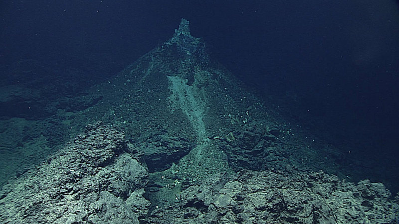 Hydrothermal chimney (white; approximately one meter tall) on top of hydrothermal mound (dark; ~10 meters tall); volcanic rocks in foreground.