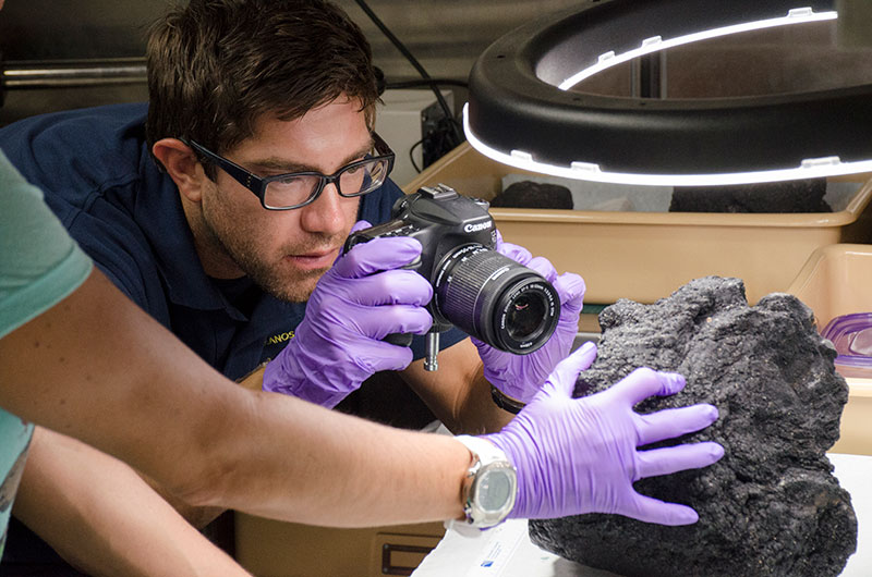 Matt Dornback, from the NOAA National Centers for Environmental Information, photographs a rock sample from the day’s dive. Rock samples are being collected during most dives for later age-dating and geochemical analysis to provide more information about the geologic history and age of seamounts in the Johnston Atoll Unit. Image courtesy of NOAA Ocean Exploration, 2017 Laulima O Ka Moana.