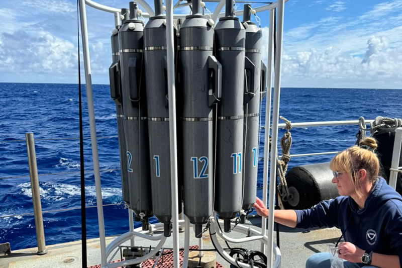 Image of the CTD and sample data manager Jennifer demonstrating how to collect the water samples from the Niskin bottles during the Beyond the Blue: Papahānaumokuākea Mapping 1 expedition. There are 12 bottles on the CTD and they are arranged in a circle, placed in chronological order.