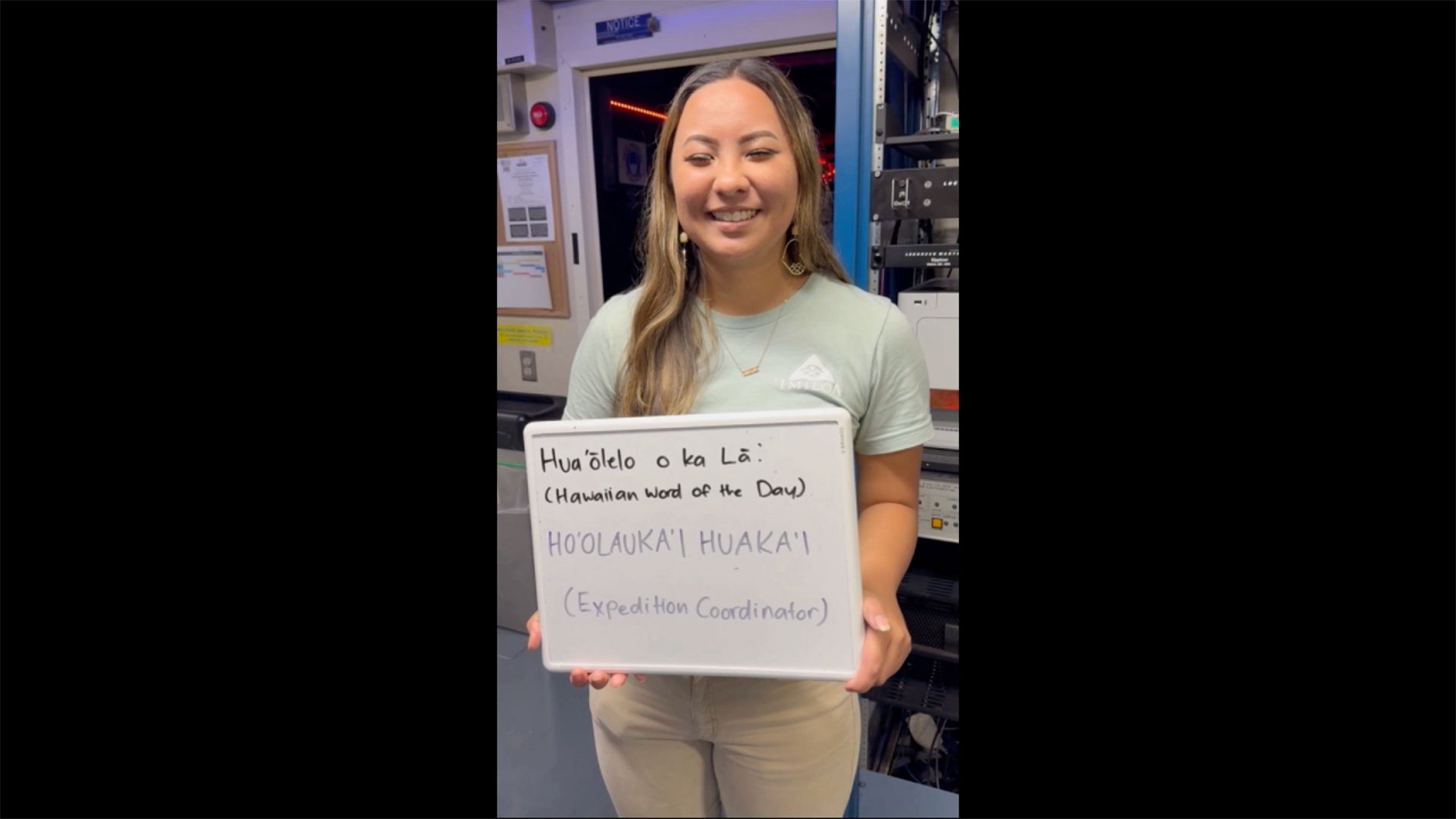 The image shows a woman standing indoors, smiling, and holding a whiteboard with handwritten text. She has long, straight hair and is wearing a light green T-shirt with a subtle logo, beige pants, and hoop earrings. The background includes equipment and posters, suggesting a professional or technical setting, possibly a control room or office. The lighting is bright, illuminating the woman's face and the text on the board clearly.