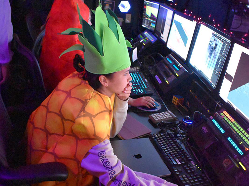 A person in a pineapple costume working at a desk with multiple computer monitors in a control room.