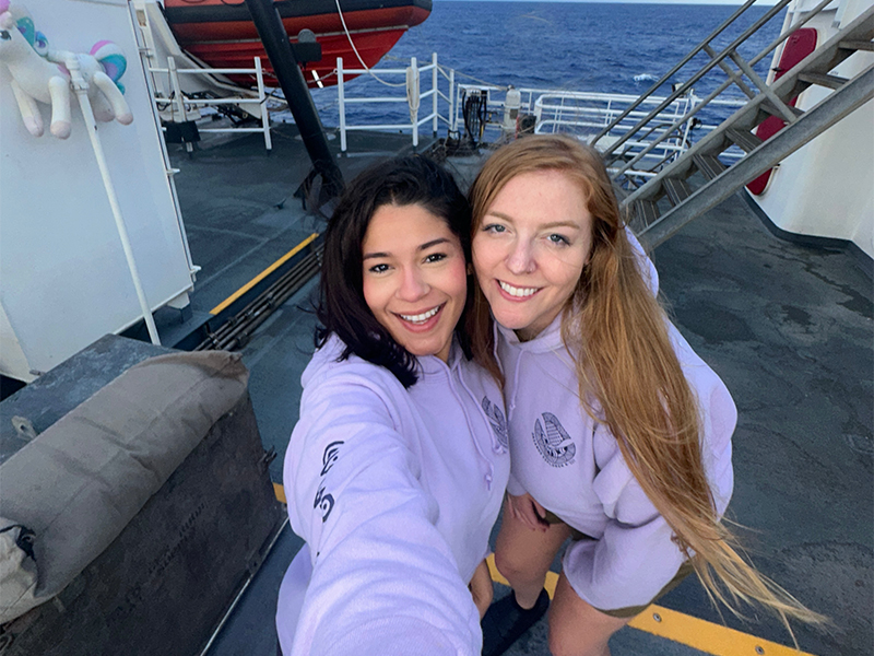 The image shows two smiling women standing on the deck of a ship, with the ocean visible in the background. Both are wearing light purple hoodies with logos and have straight hair; one has dark hair, and the other has light brown hair. Behind them, a red inflatable boat is mounted, and a set of stairs leads up to another level of the ship. To the left, a white container with a plush toy, resembling a unicorn, is partially visible. The decking is dark grey, with yellow and white lines marking pathways.