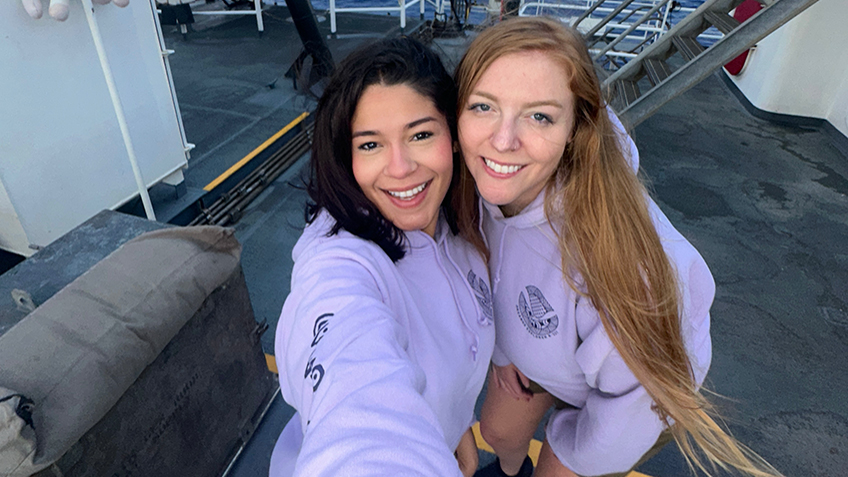 The image shows two smiling women standing on the deck of a ship, with the ocean visible in the background. Both are wearing light purple hoodies with logos and have straight hair; one has dark hair, and the other has light brown hair. Behind them, a red inflatable boat is mounted, and a set of stairs leads up to another level of the ship. To the left, a white container with a plush toy, resembling a unicorn, is partially visible. The decking is dark grey, with yellow and white lines marking pathways.