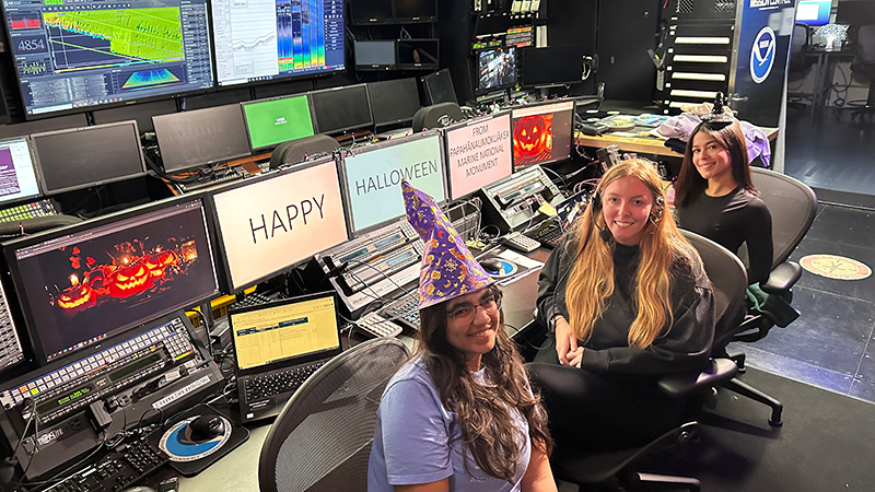 Three women in a control room, two of them wearing witch hats, surrounded by monitors displaying Halloween decorations.