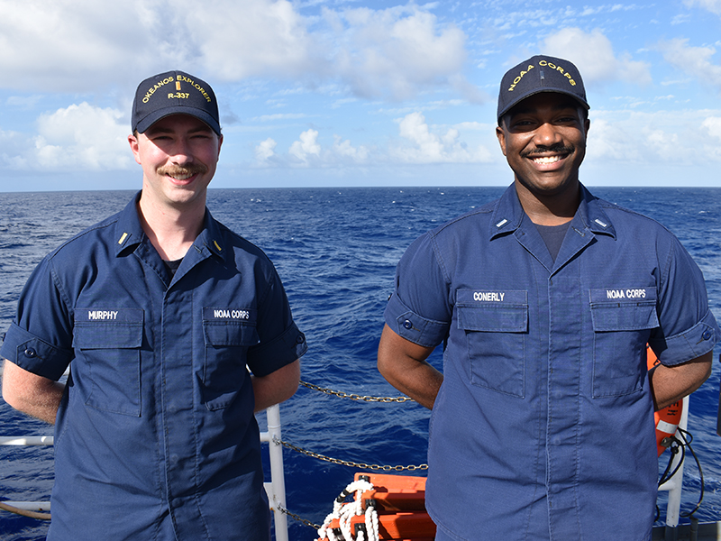 Two NOAA Corps officers standing on a ship's deck with the ocean in the background.