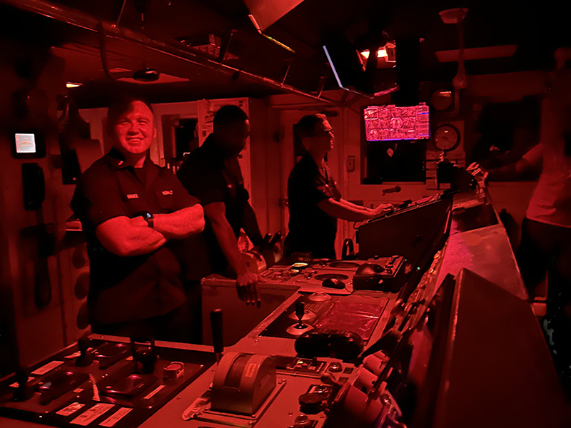 Four people in a dimly lit ship control room under red lighting, engaging with control consoles and instruments.