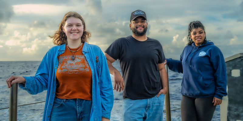 Three people standing on a vessel, with the sea and cloudy sky in the background.