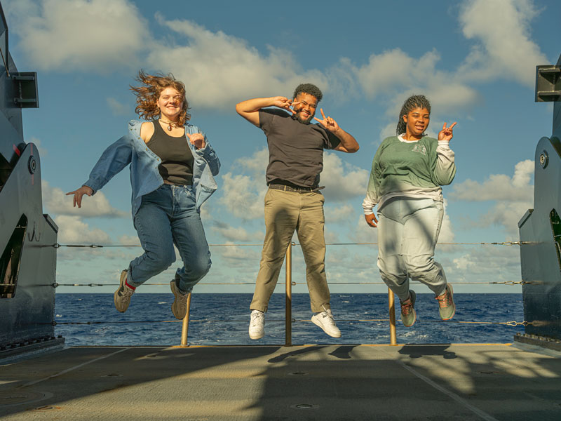 Explorers-in-Training (from left to right) Abby Norstad,  Nathanael Mathis, and Gina Knox jump for joy on the back deck of NOAA Ship Okeanos Explorer, demonstrating their enthusiasm for their at-sea experience with NOAA Ocean Exploration.