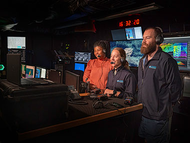 Three people in a control room with multiple monitors, wearing headsets.