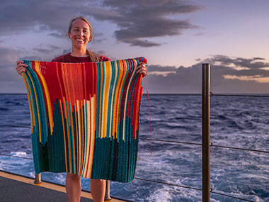 Image of NOAA Ocean Exploration Internship Program Coordinator Jesse Gwinn on the fantail of NOAA Ship Okeanos Explorer with her in-progress bathymetry blanket, which illustrates ocean depth data collected during the Beyond the Blue: Johnston Atoll Mapping 2 expedition.