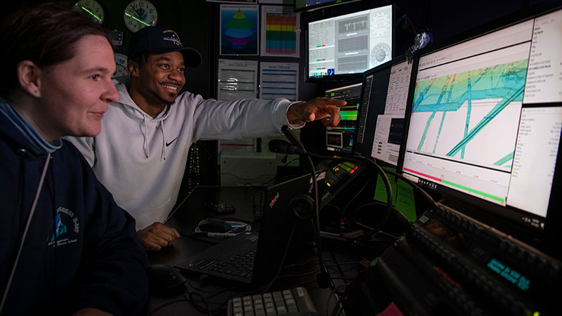 Two people at a control station interacting with computer screens displaying data visualizations.